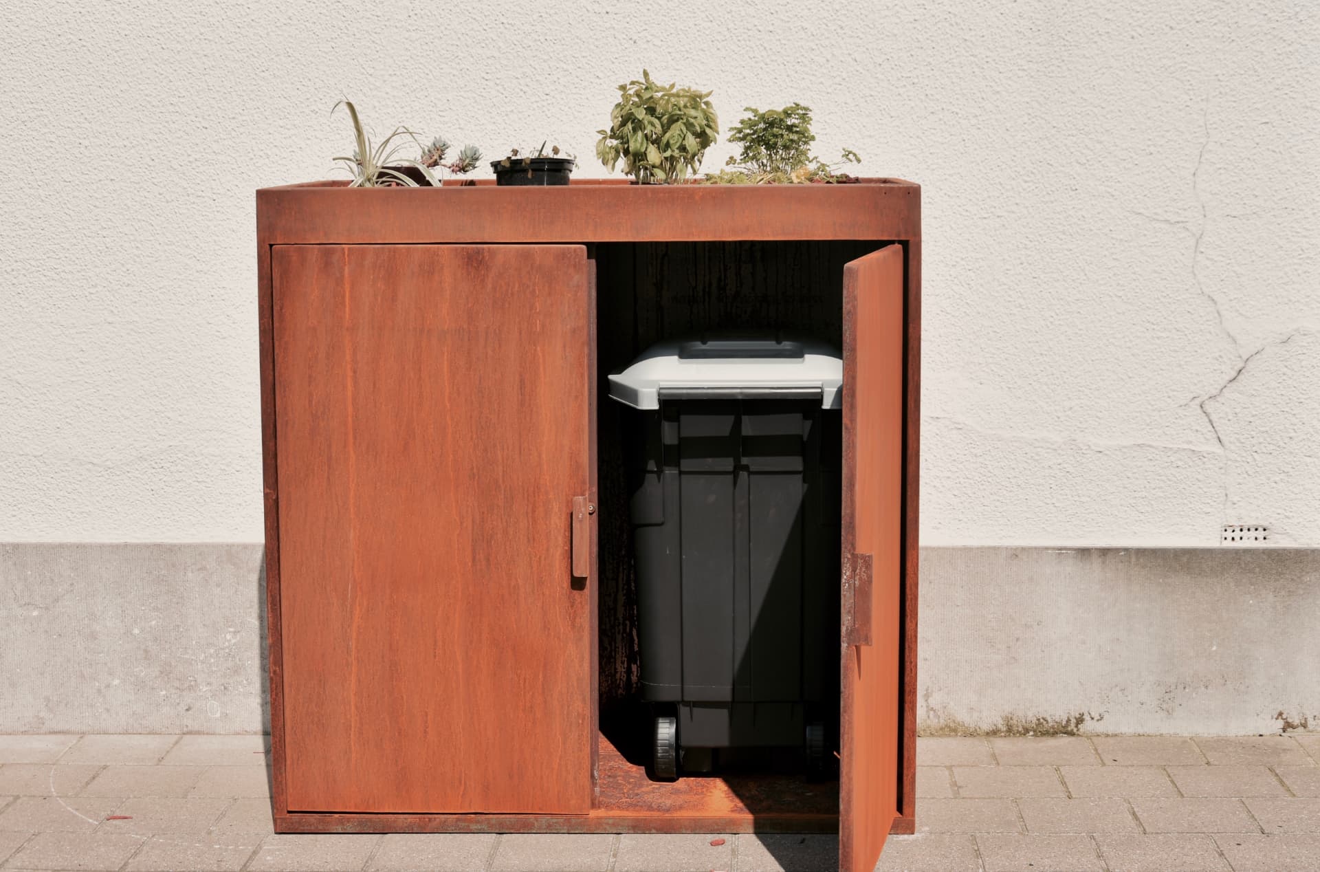 KUJP Kliko in cortenstaal met beplante bovenkant en een zichtbare rolcontainer achter de open deur